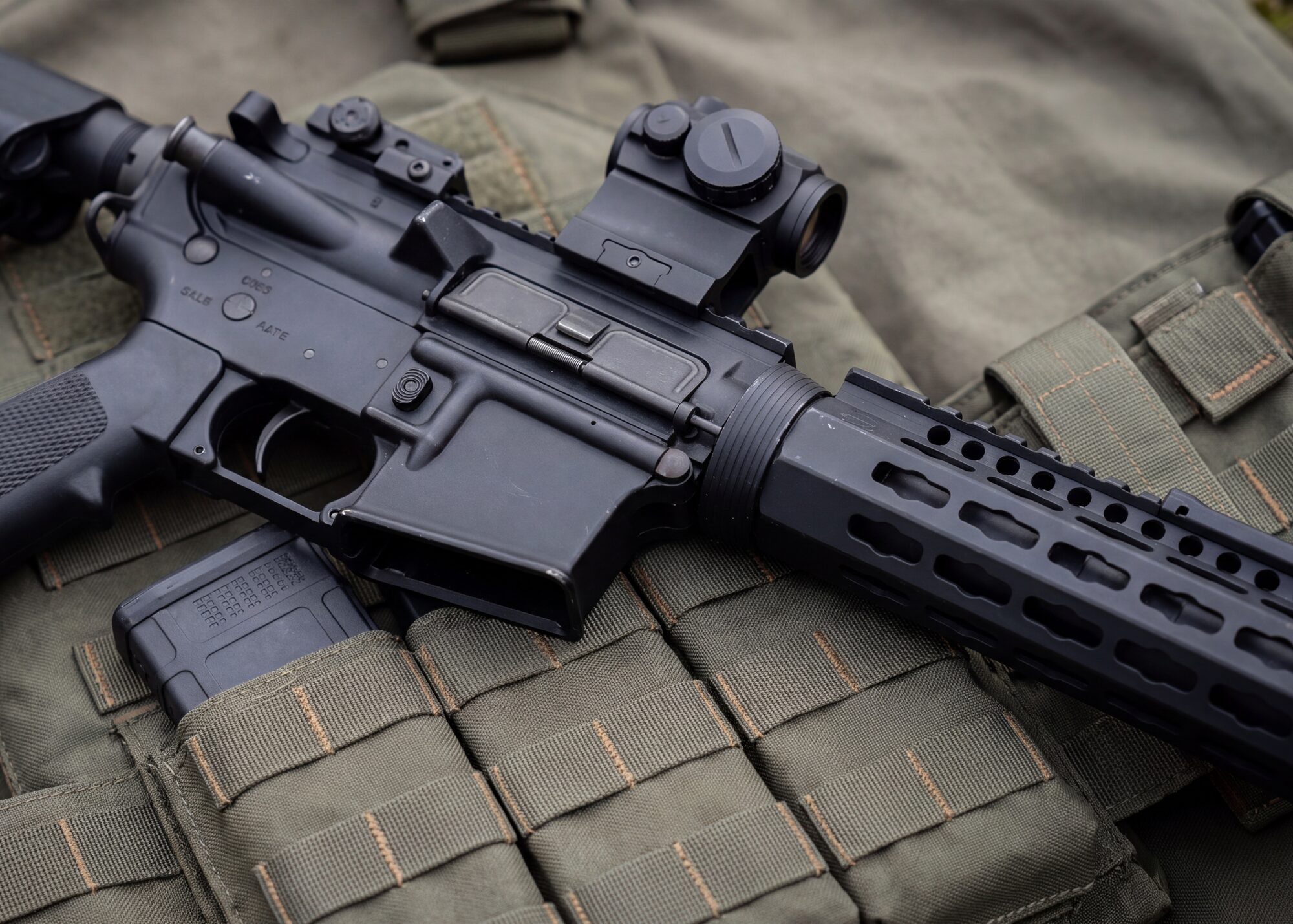 Mitra elettrico softair tipo M4 con ottica red-dot appoggiato sulla fettuccia MOLLE verde di un corpetto tattico
